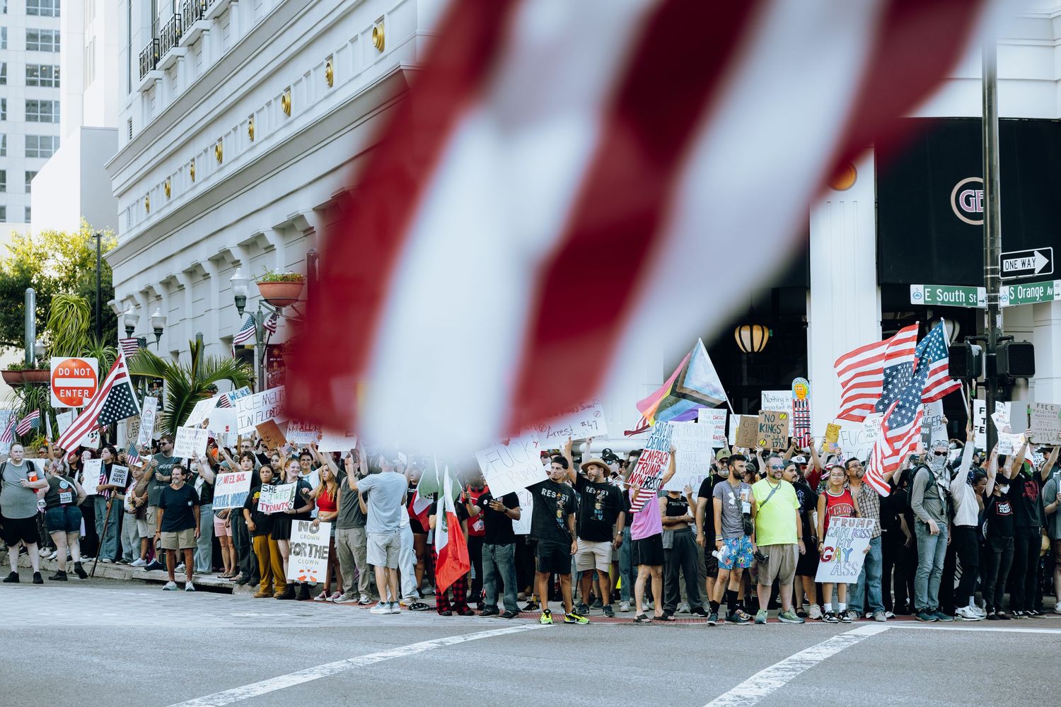 A large crowd of protesters line a street with American flags and buildings visible in the background.