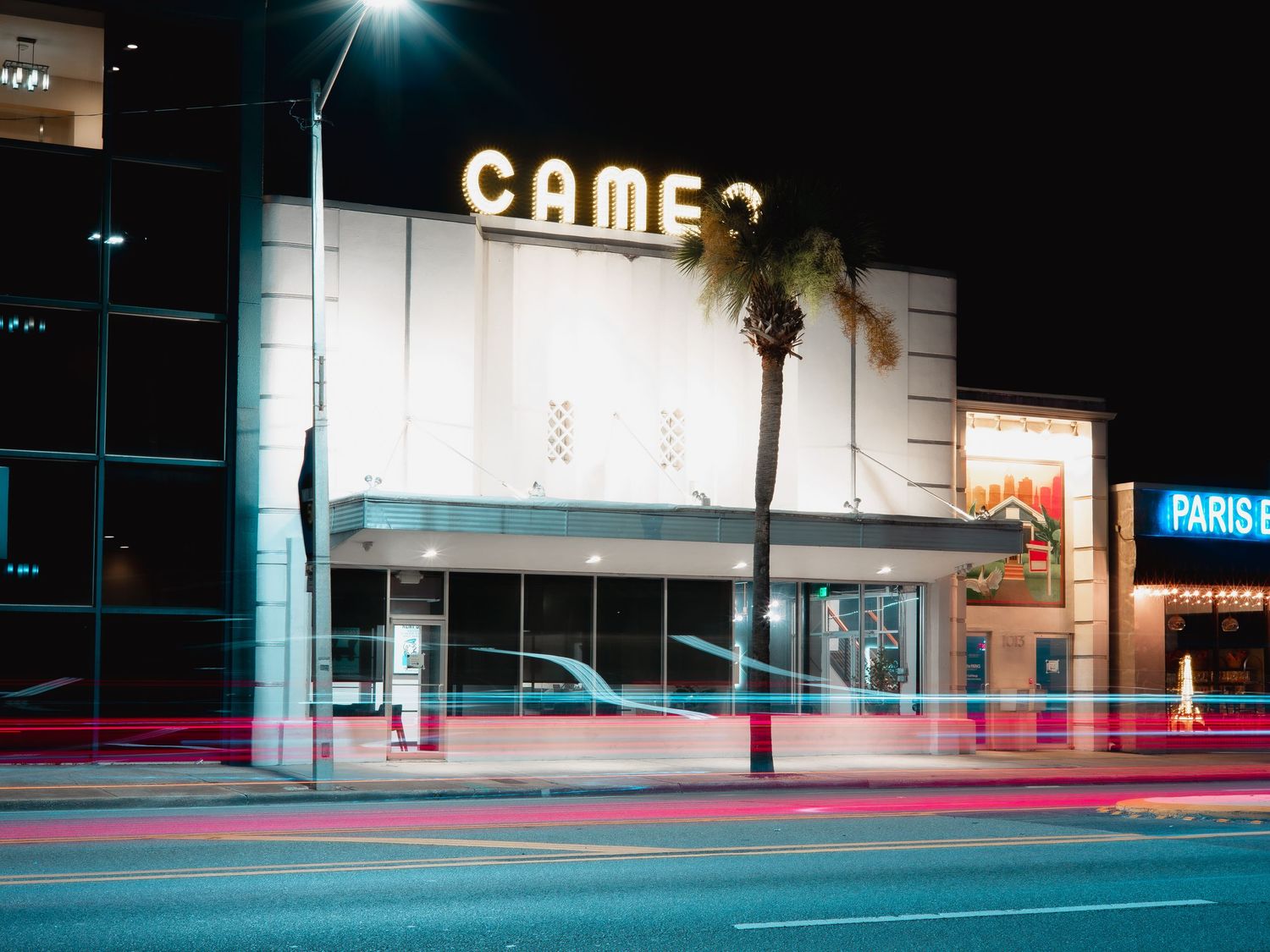 Long exposure photograph shows light trails and illuminated storefronts at night in urban setting.