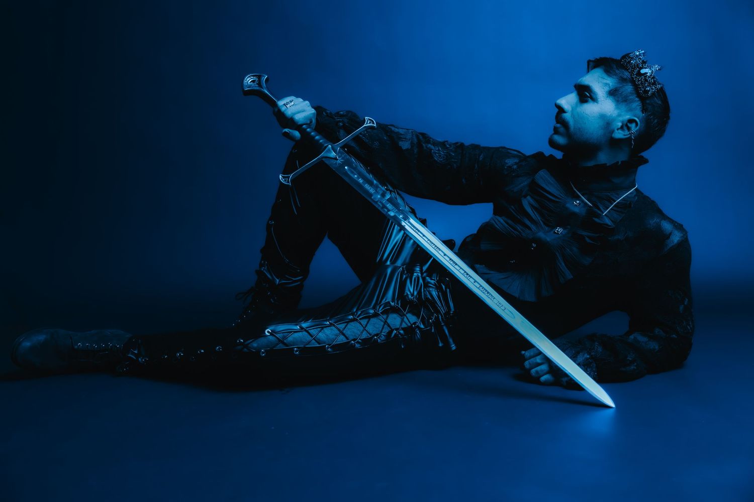 Man in black leather outfit poses dramatically on floor holding a medieval sword in blue-lit studio setting.