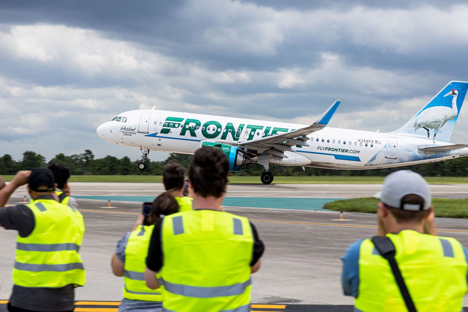 Frontier Airlines Airbus A320 aircraft taxiing on runway while ground crew in yellow vests watch and photograph.