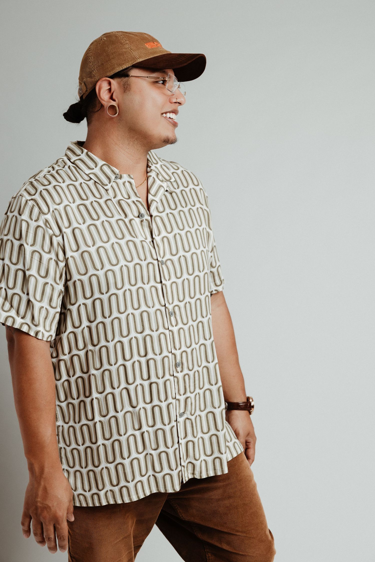 Man smiling in patterned cream short-sleeve shirt, brown corduroy pants, and tan cap against gray background.