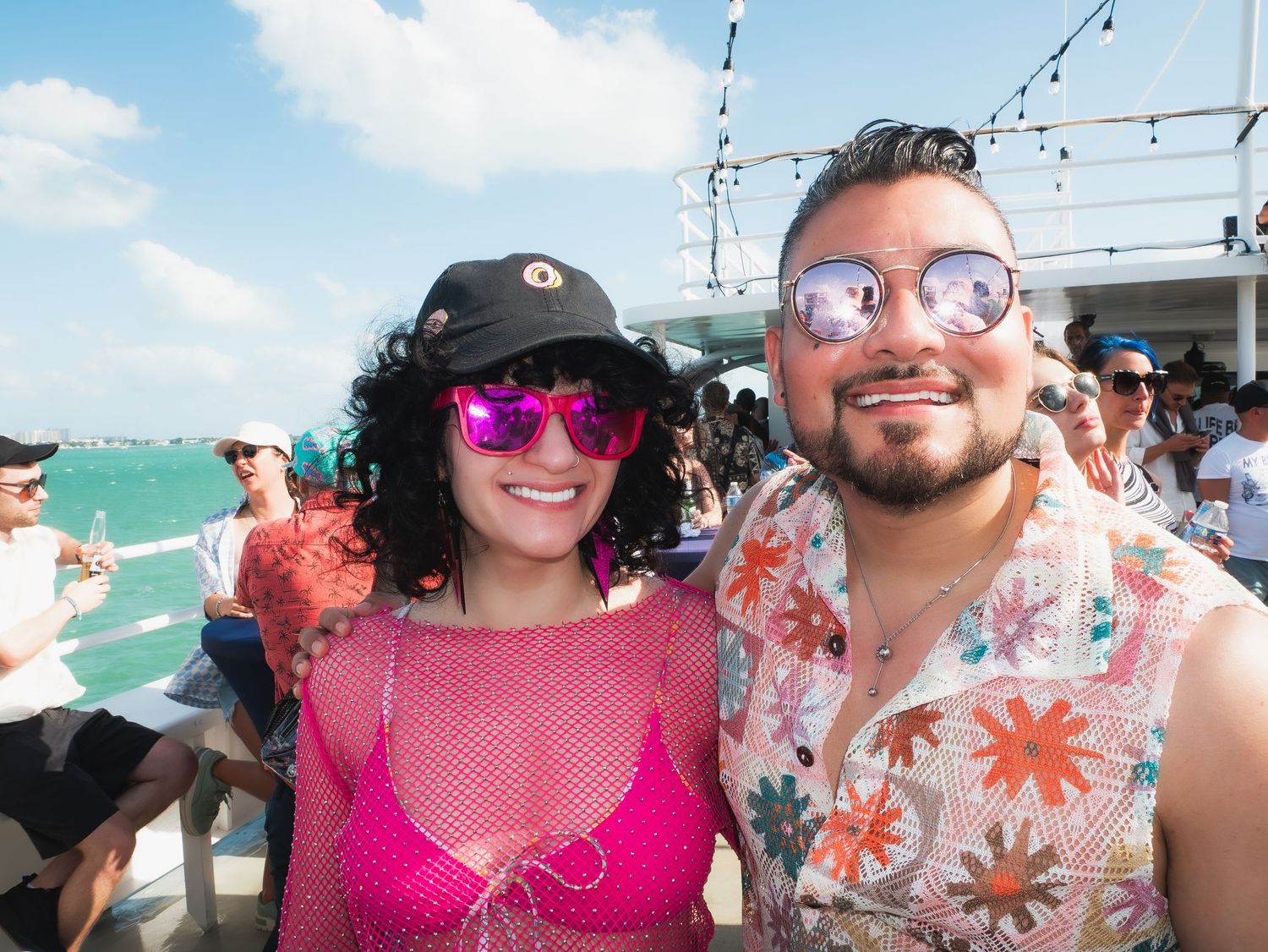 Two people wearing sunglasses smile together on a boat deck during a sunny day at sea, dressed in summer attire.