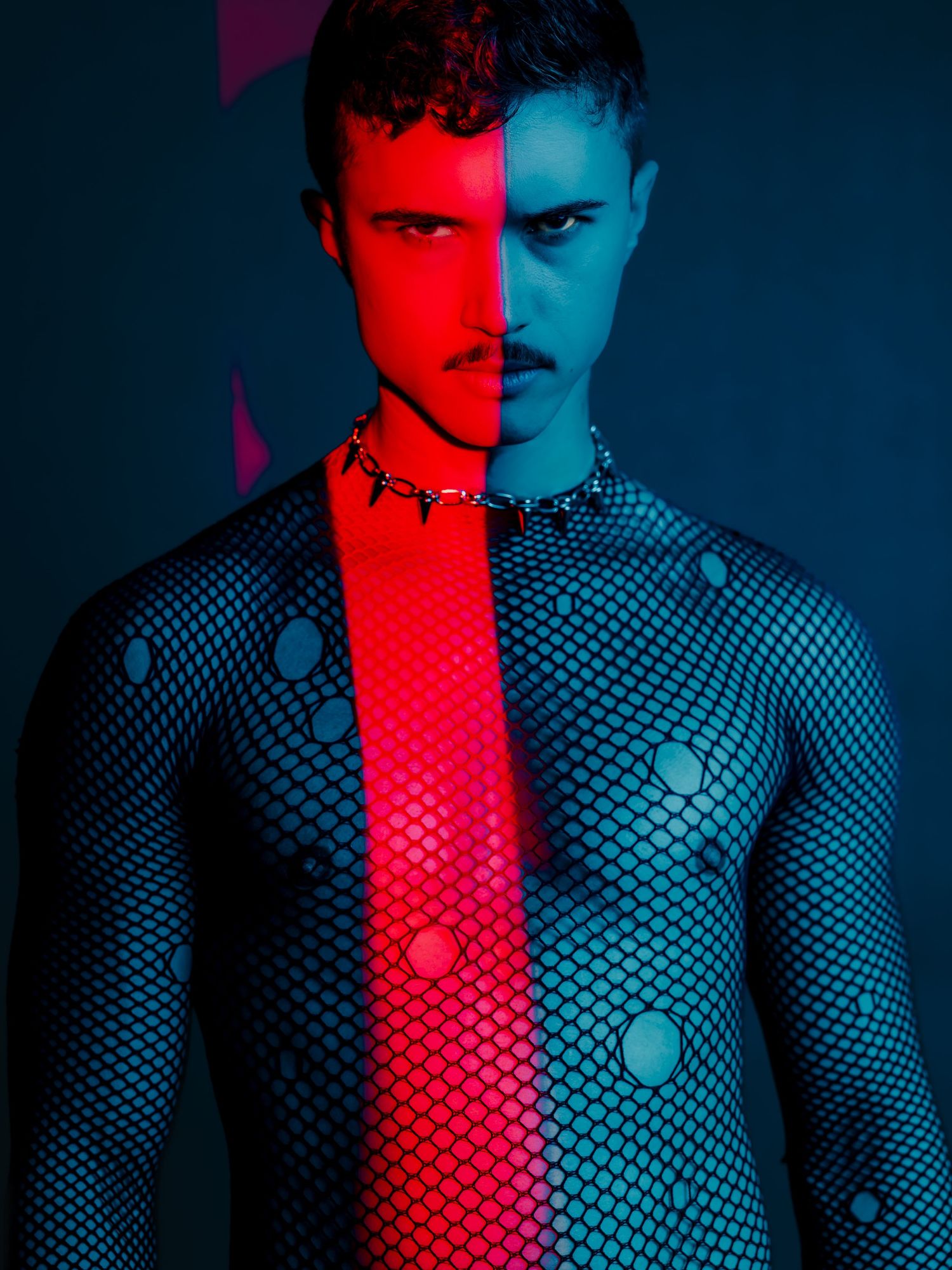 Man in fishnet bodysuit with polka dots under dramatic red and blue split lighting wearing a spiked choker necklace.
