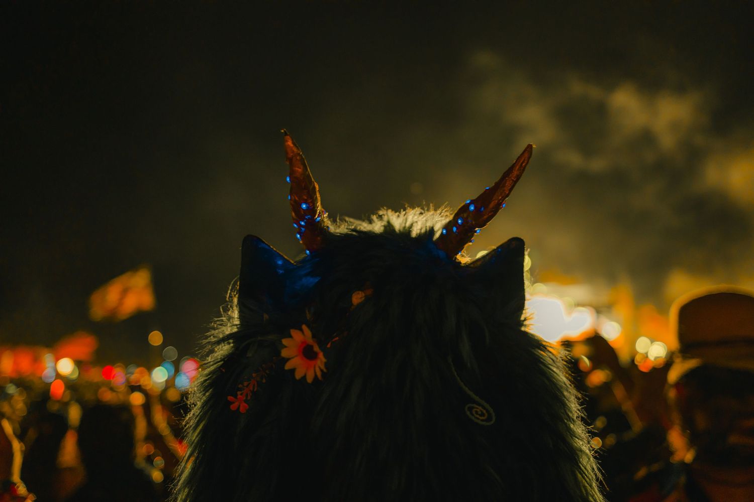A silhouetted figure with horns and flowers against a moody sunset sky at a festival with glowing lights in background.