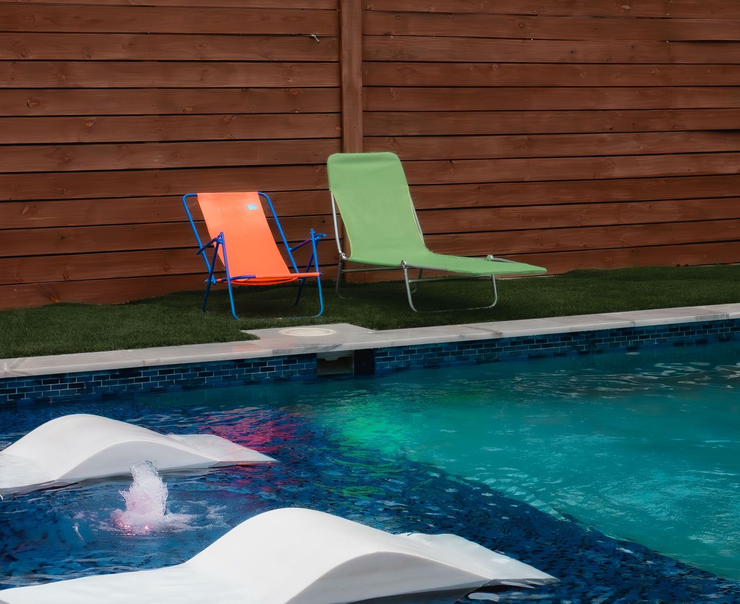 Two colorful lounge chairs, orange and green, positioned by the edge of a swimming pool with floating white rafts.
