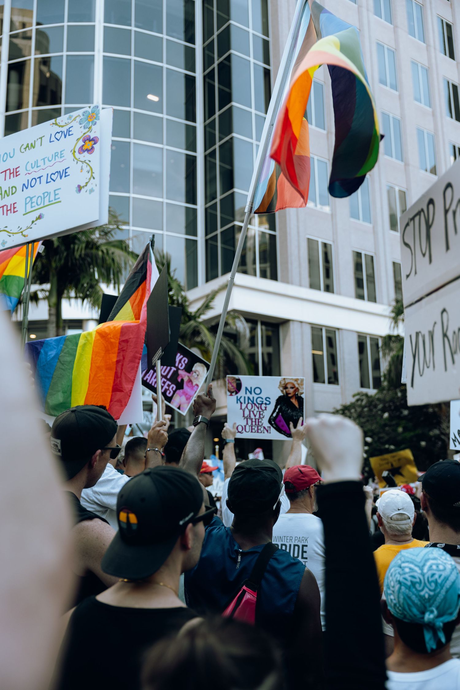 Crowd of protesters holding rainbow pride flags and signs gather outside a modern office building.
