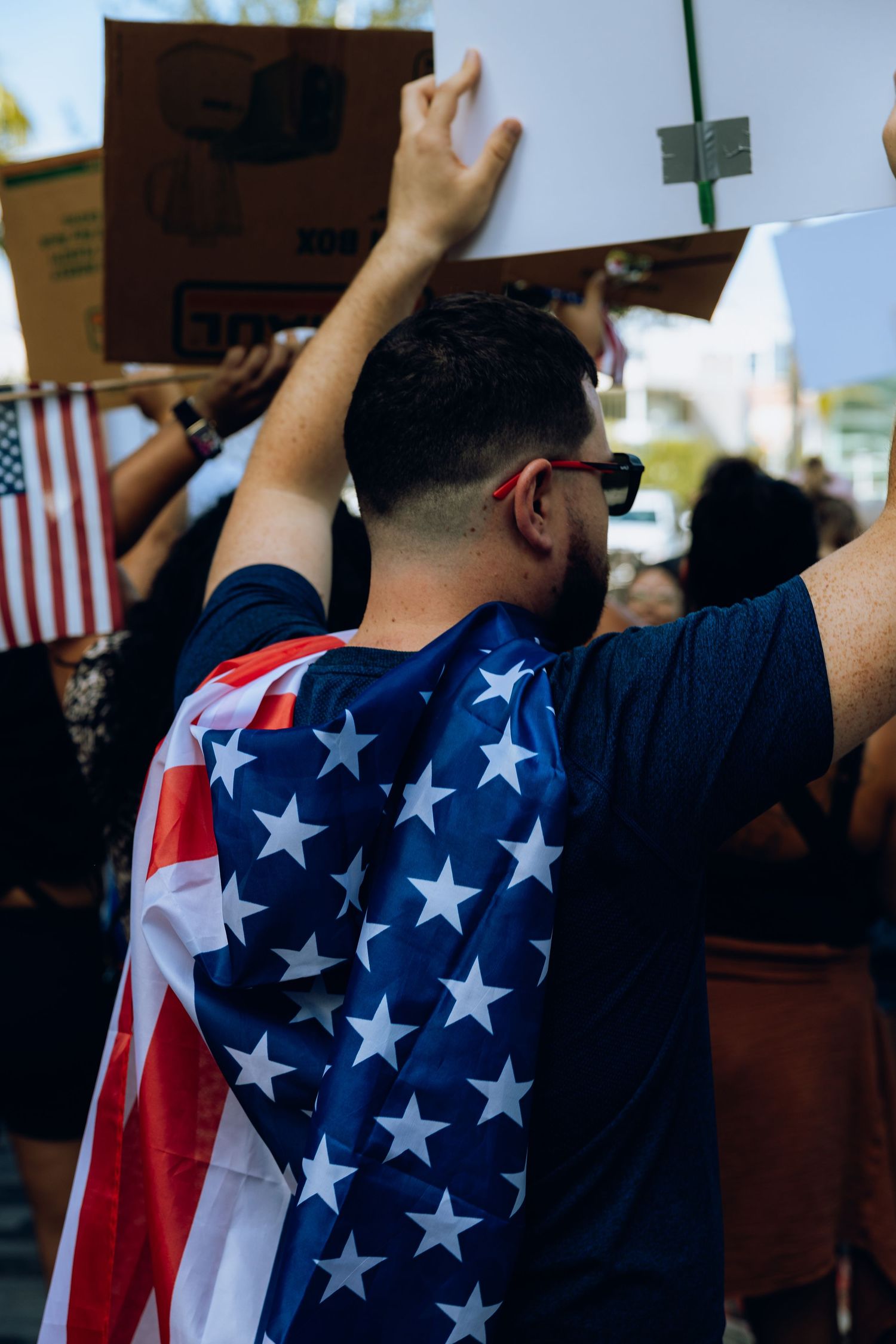 Person wearing American flag as cape participates in protest with raised hands.
