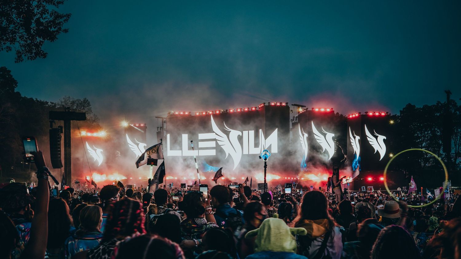 Panoramic view of a crowded outdoor music festival with red stage lighting illuminating the night sky.