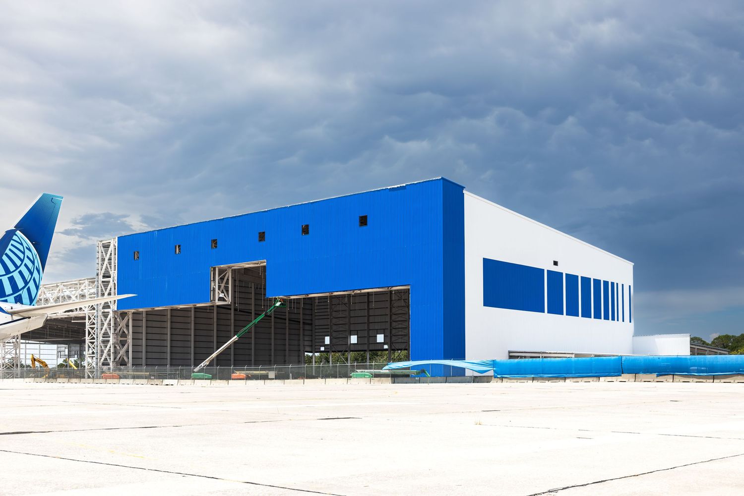 Large blue aircraft maintenance hangar at an airport under cloudy skies with a United Airlines plane visible.