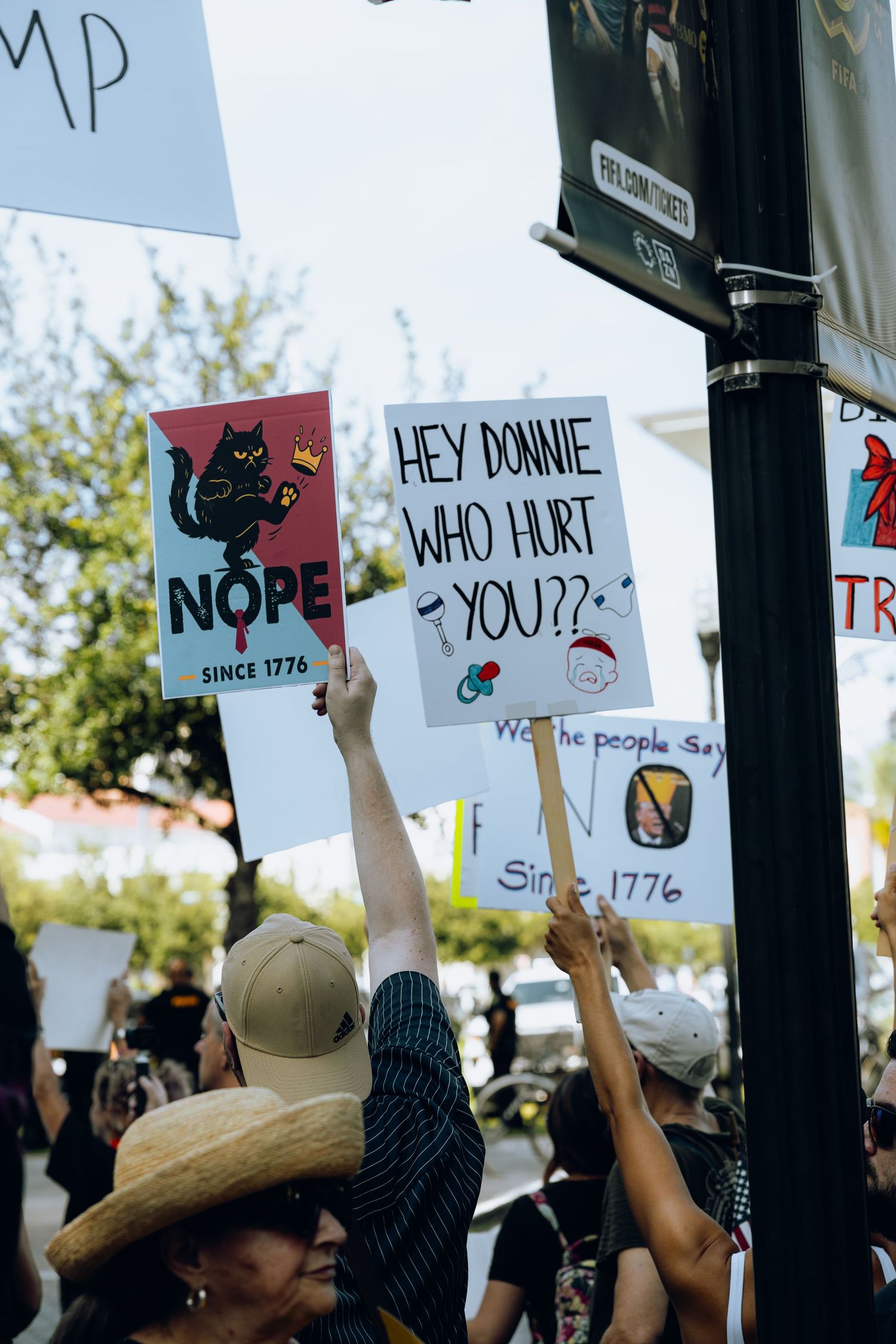 Protestors at a demonstration hold up various handmade signs against a bright sunny sky in an outdoor gathering.