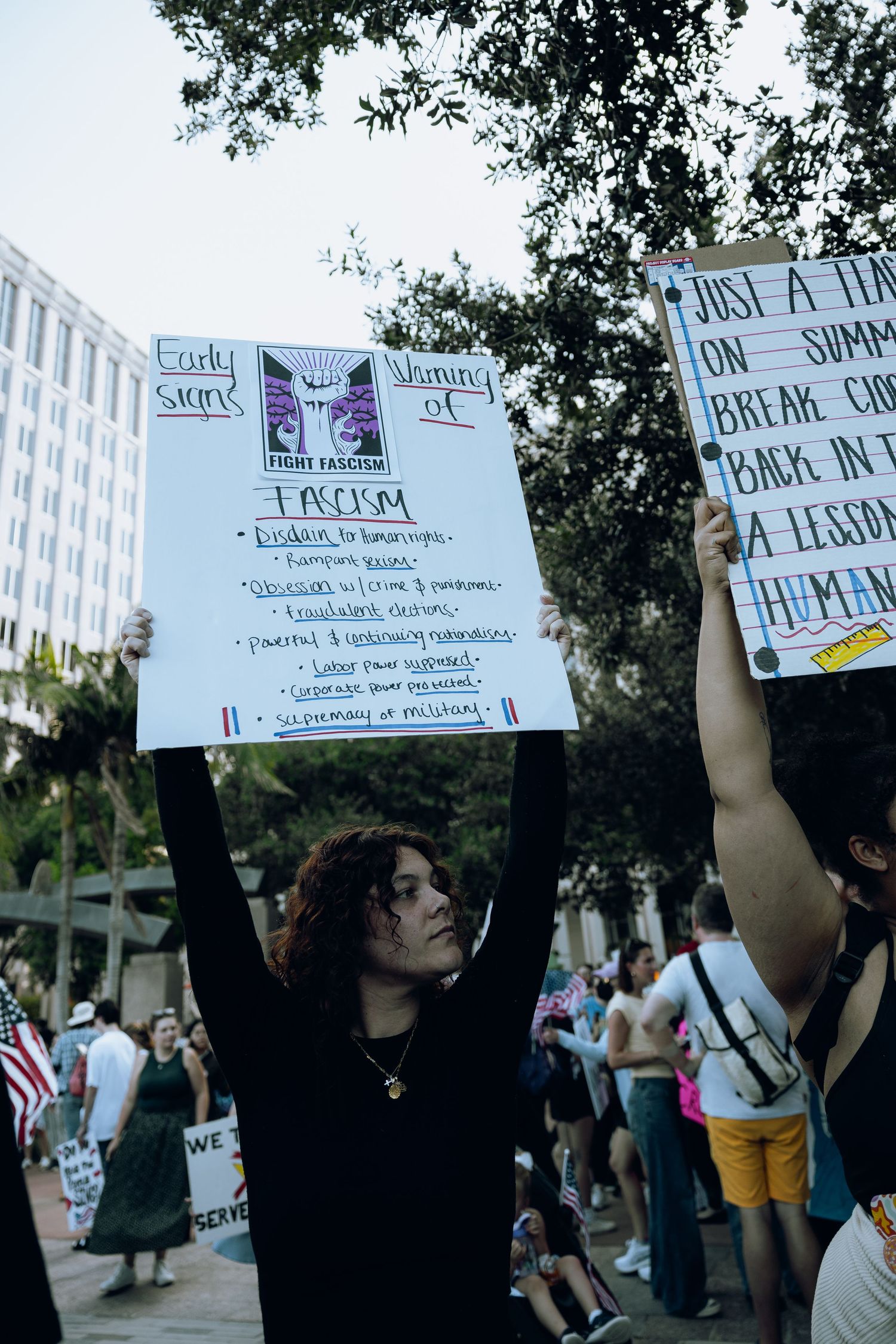 Person in black holds protest sign at demonstration with urban buildings in background.