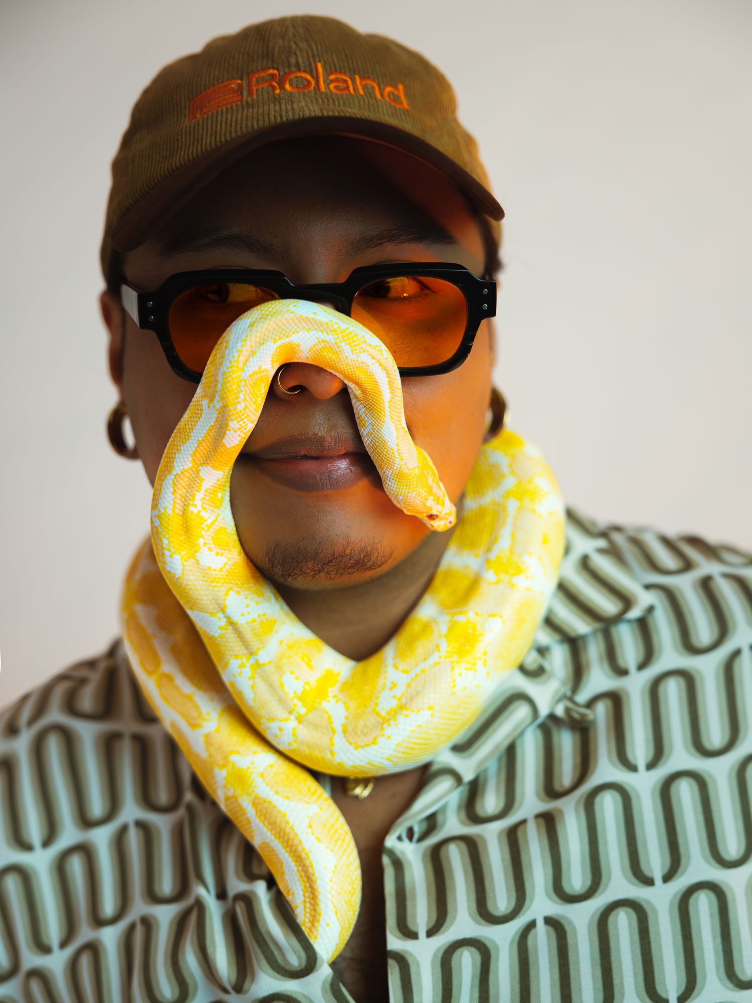 A person wearing orange sunglasses and a Roland cap poses with a yellow albino python draped around their face and neck.