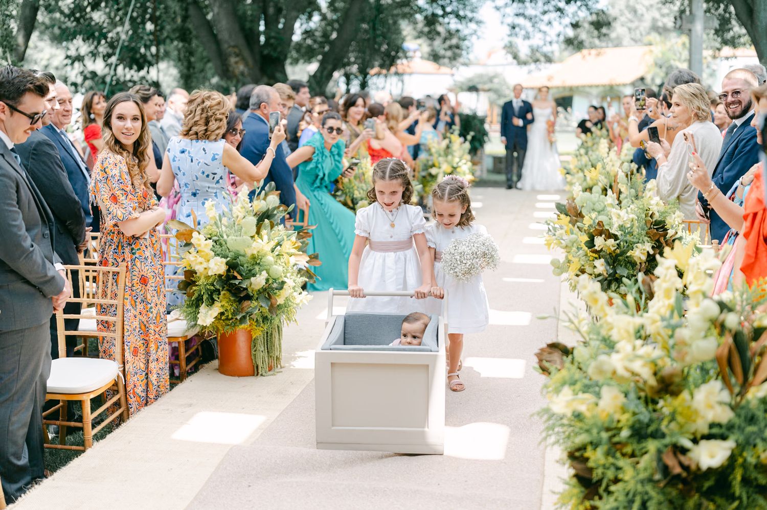 Flower girls push a baby in a decorative cart down the wedding ceremony aisle lined with flowers.