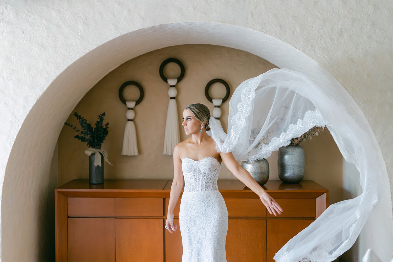 Bride in a strapless lace wedding gown with flowing veil under an elegant arched doorway.
