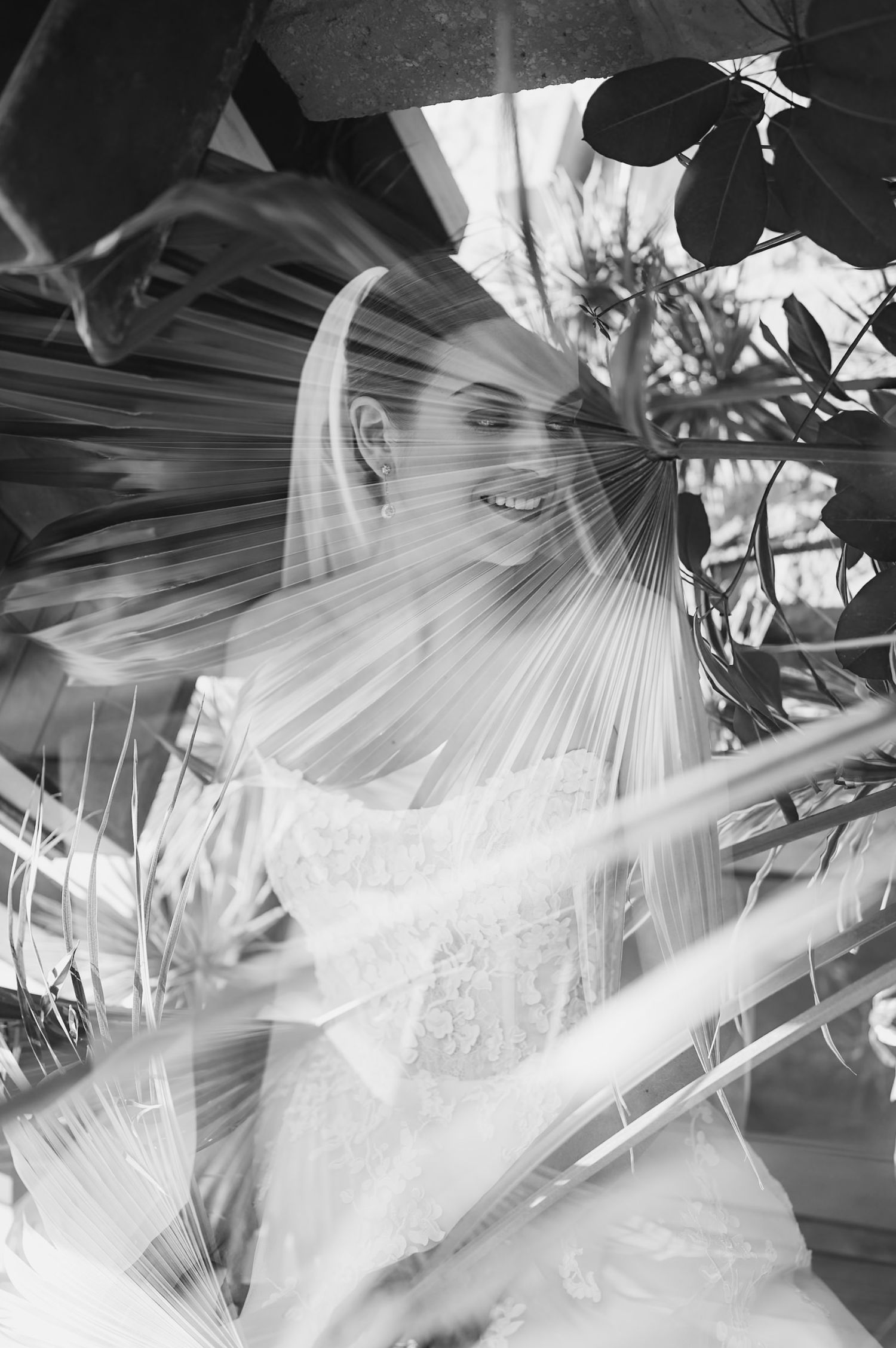 Artistic black and white portrait of smiling bride in veil framed by tropical palm leaves.