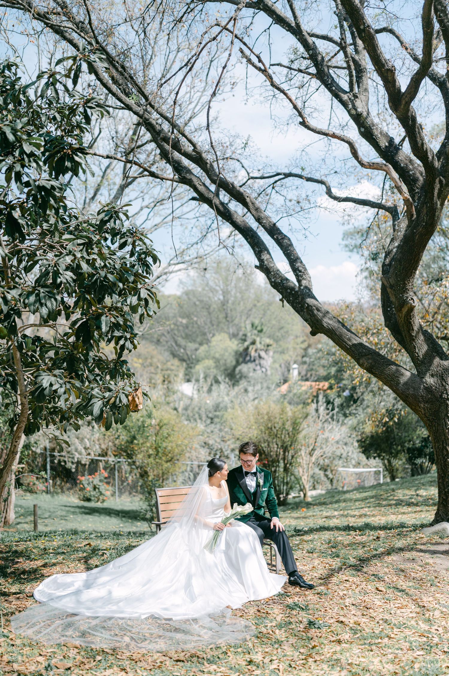 Bride and groom sit together on wooden bench in sunlit garden, bride in ball gown with long veil, groom in green suit.