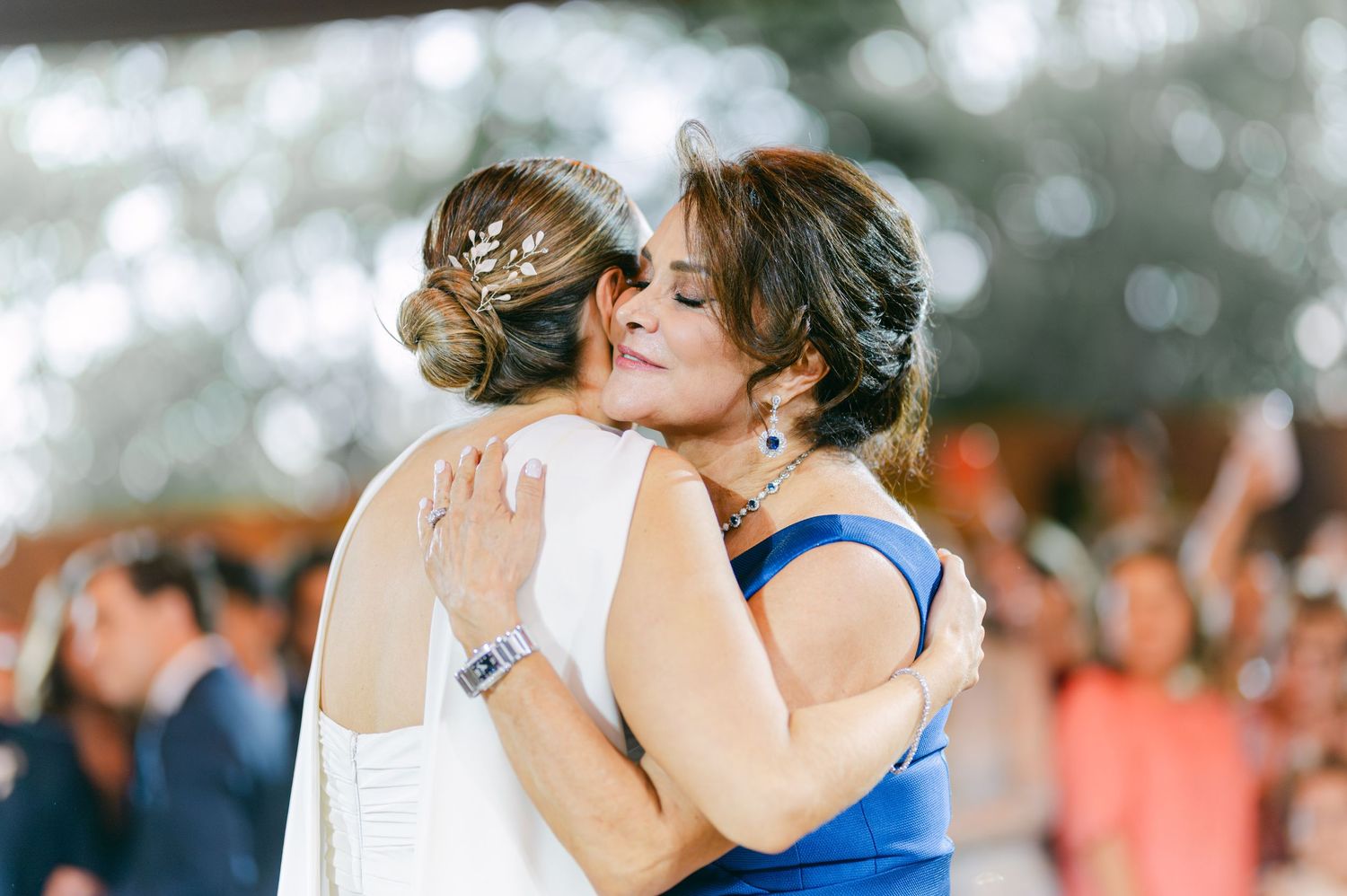 Bride in white gown shares a heartfelt hug with a woman in blue dress during wedding celebration.