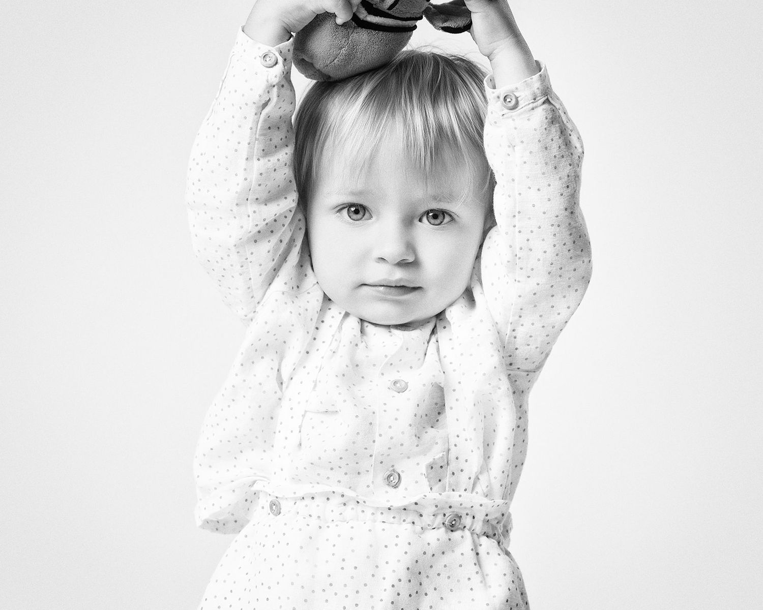 Cute blonde toddler in white dotted outfit holds shoes above her head in black and white portrait.