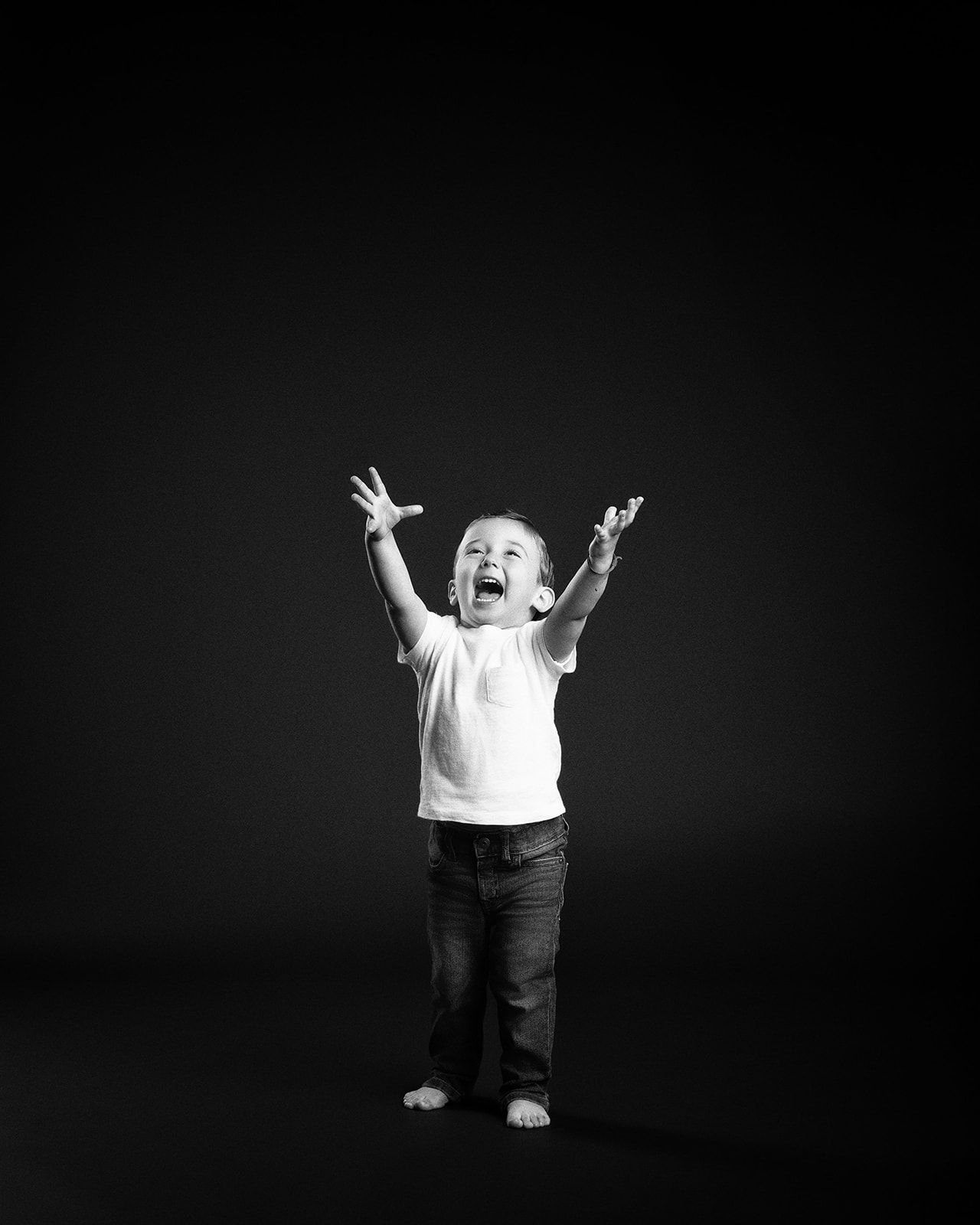 Joyful toddler boy raises arms up high, wearing white t-shirt and jeans in black and white studio photo.
