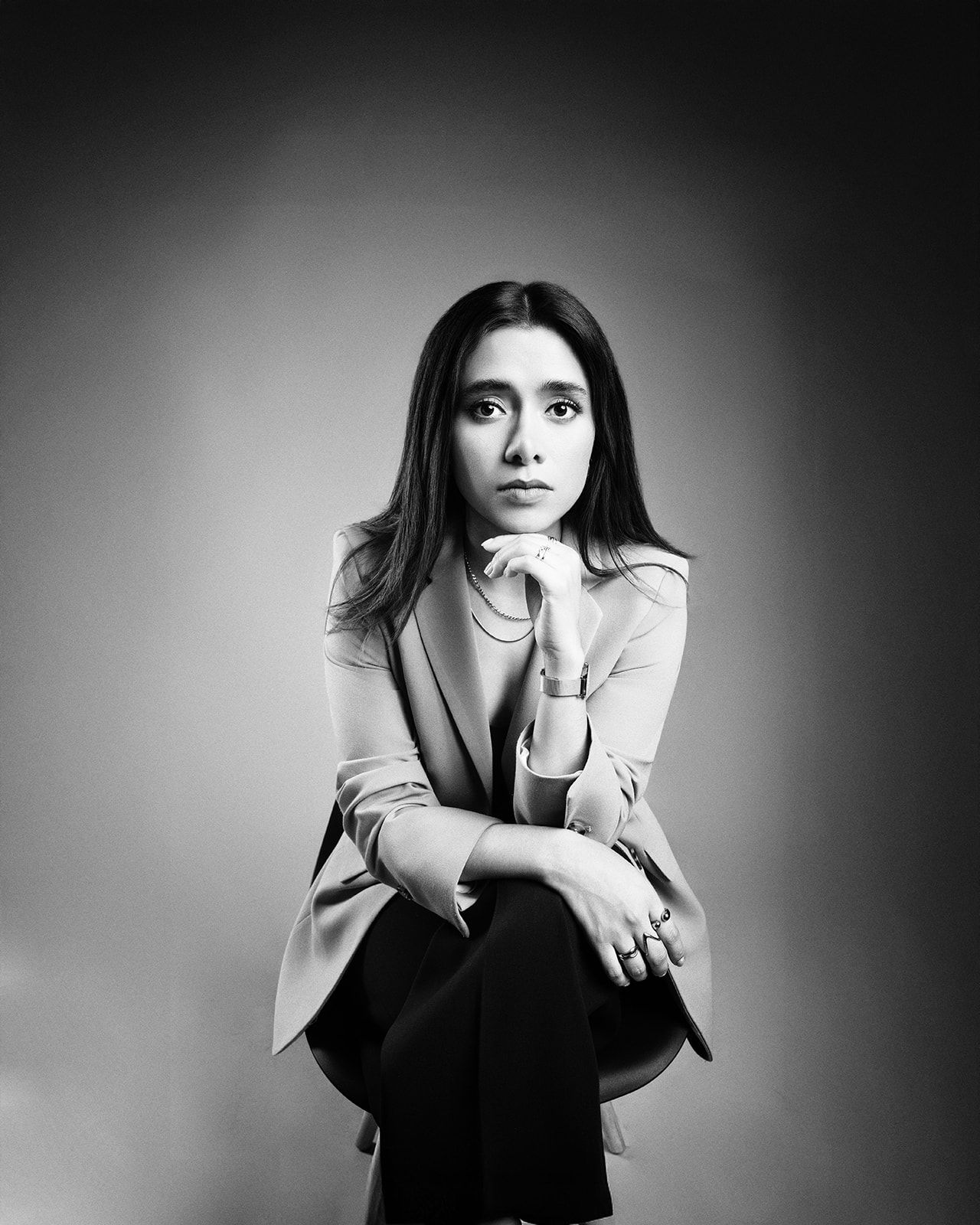 Black and white portrait of a young woman in a blazer, sitting pensively with hands clasped under her chin.