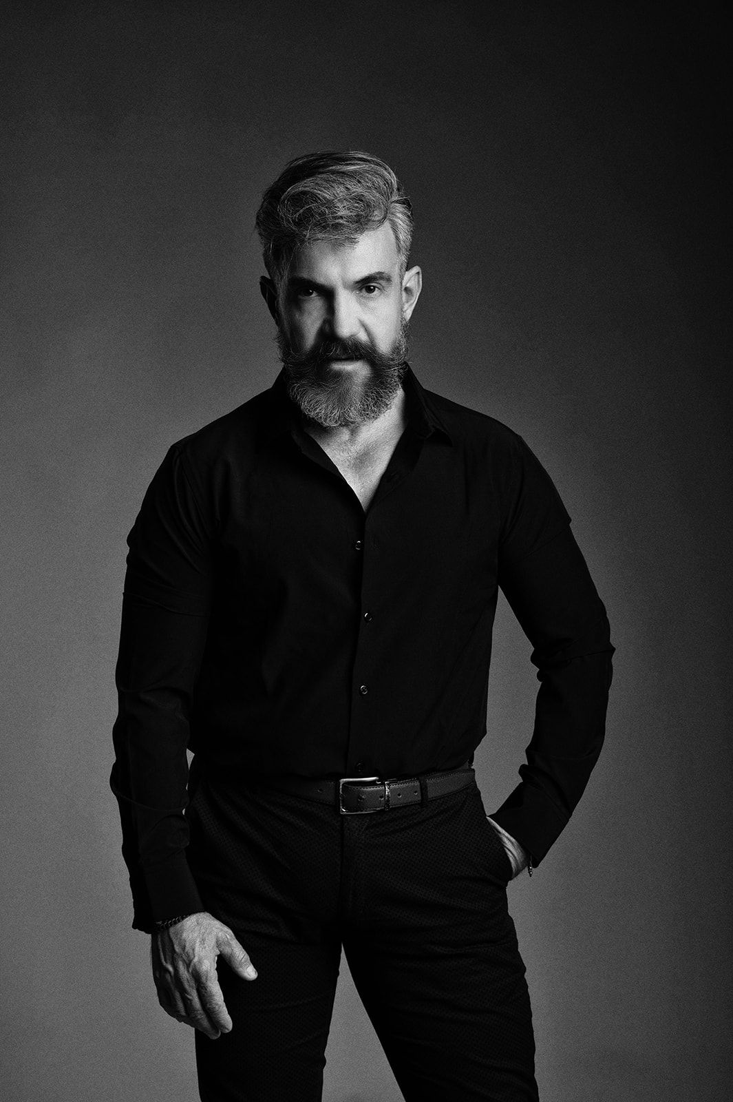 Black and white photo of bearded man in black shirt and pants posing confidently.