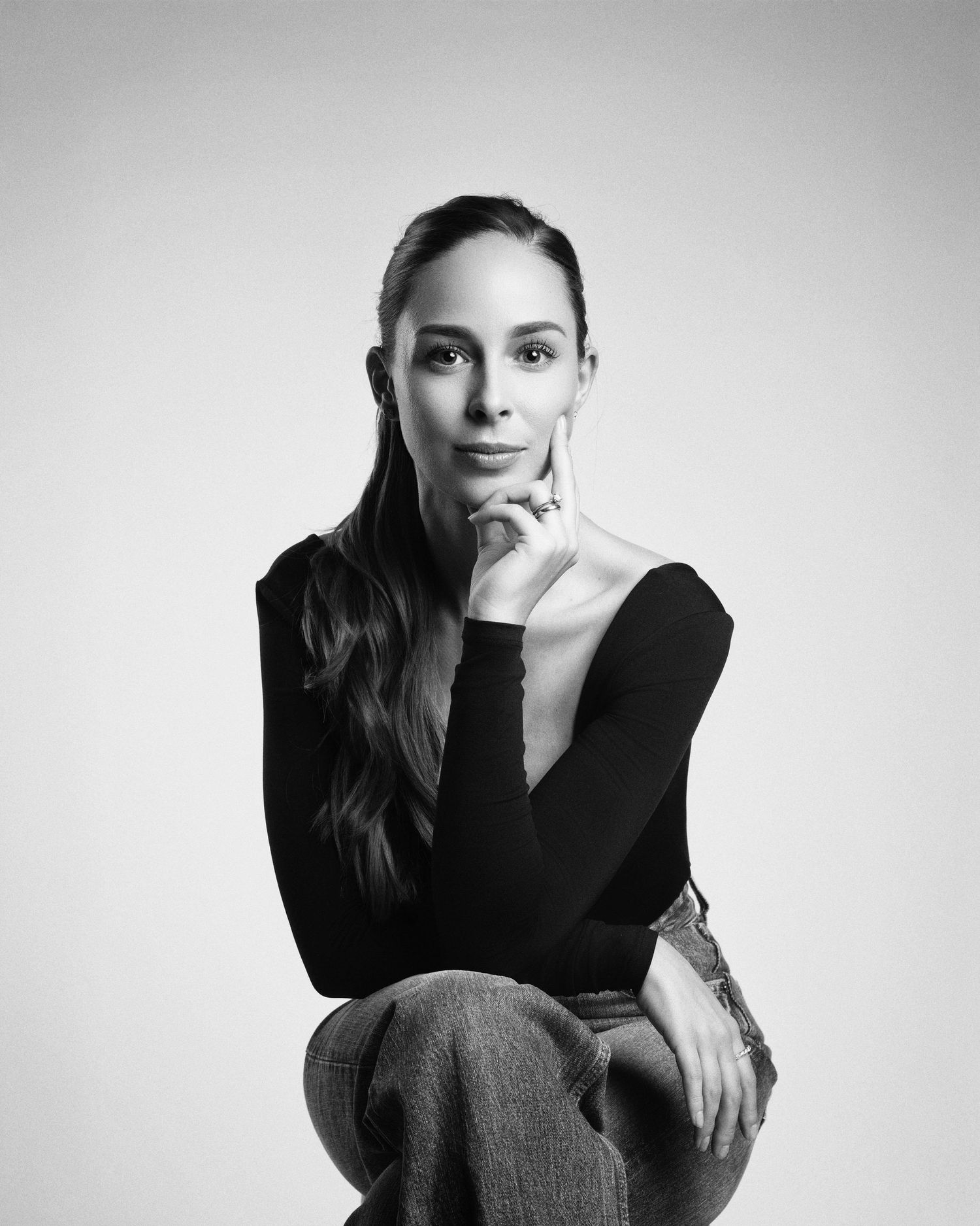 Black and white studio portrait of a young woman in black long-sleeve top and jeans, hand on chin.