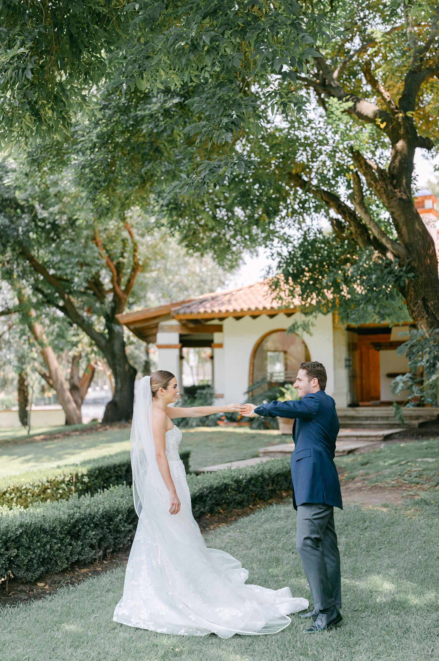 Groom in navy suit reaching out to bride in lace gown during outdoor first look moment.