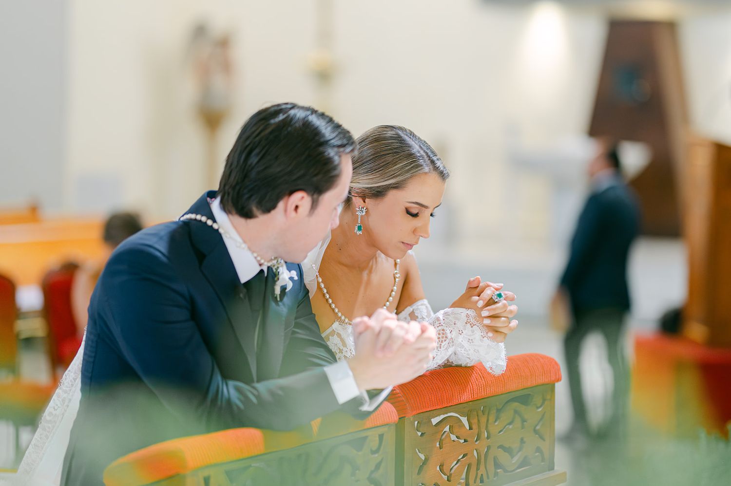 Bride and groom share intimate prayer moment together in ornate wooden church pew during wedding ceremony.