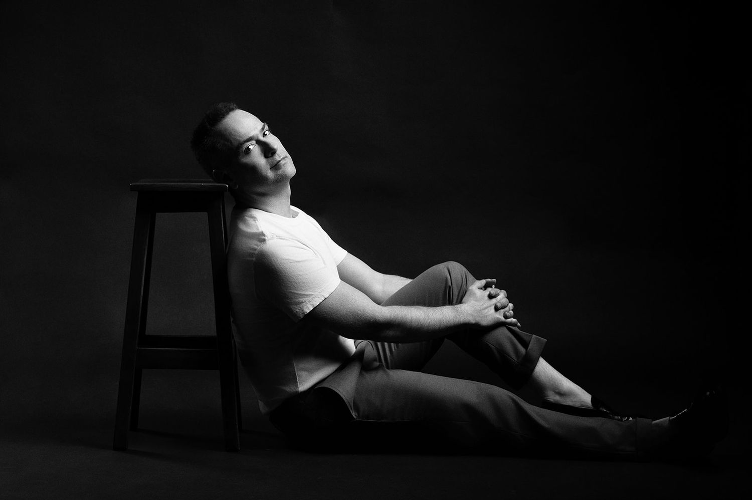 Black and white photo of a man relaxing on the floor leaning against a stool in a dark studio.