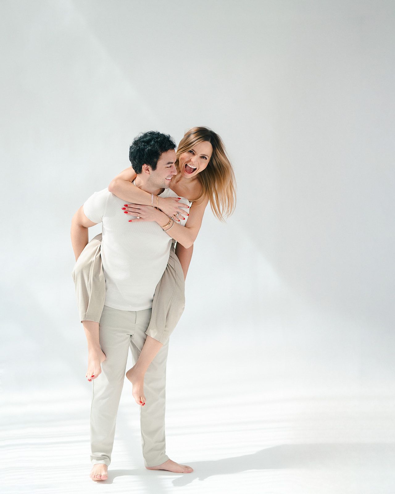 A couple sharing a joyful piggyback ride in a bright white studio, dressed in casual neutral outfits.