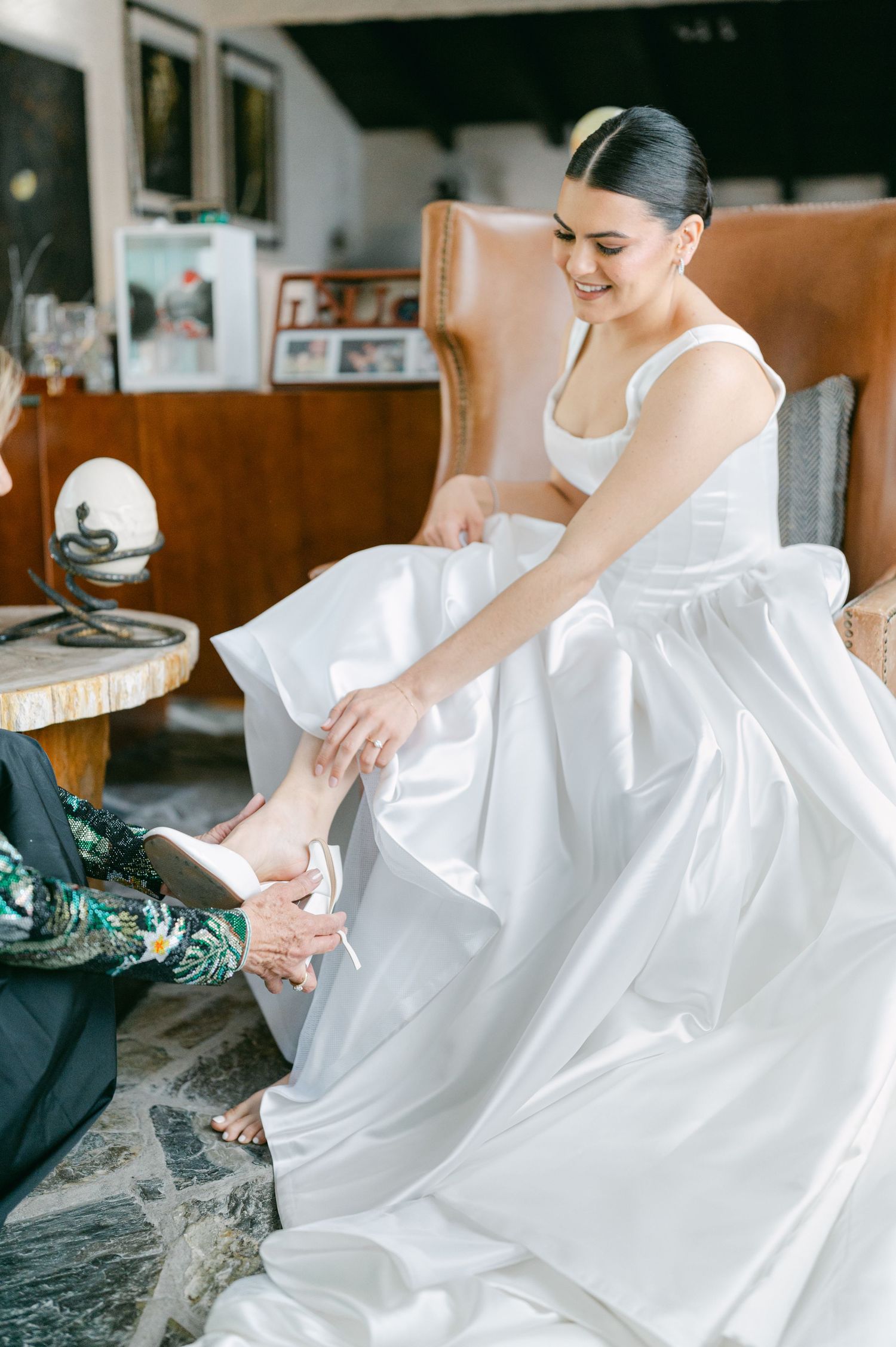 Smiling bride in white ball gown sits in leather chair while someone helps her put on her wedding shoes.