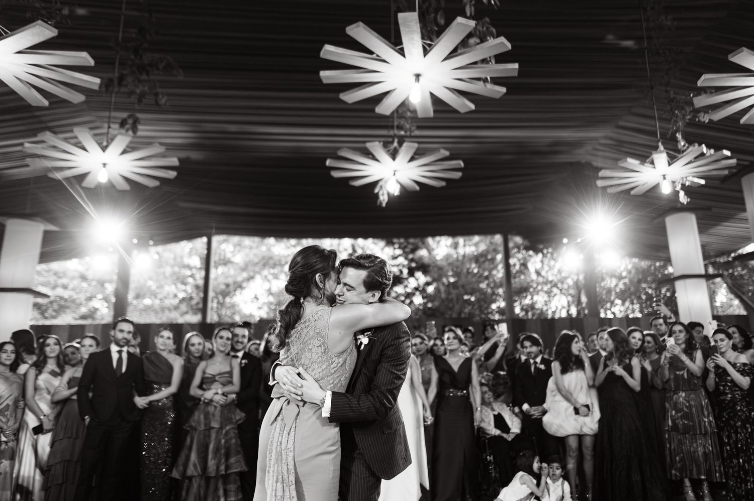 Black and white photo of newlyweds sharing first dance embrace surrounded by cheering guests at reception.