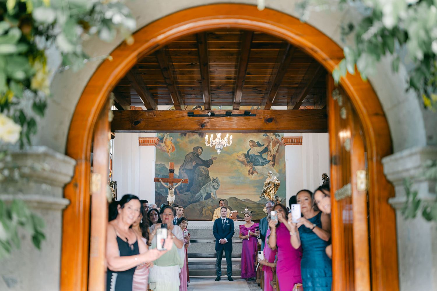 Wedding party stands beneath wooden archways decorated with greenery and flowers during an outdoor ceremony.