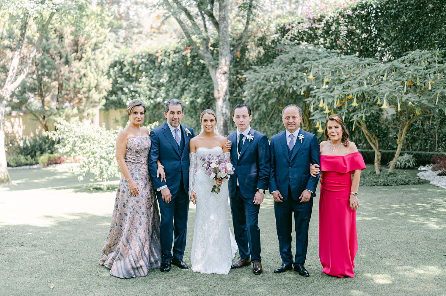 Bride and groom pose with four family members in garden, wearing navy suits and elegant formal gowns.
