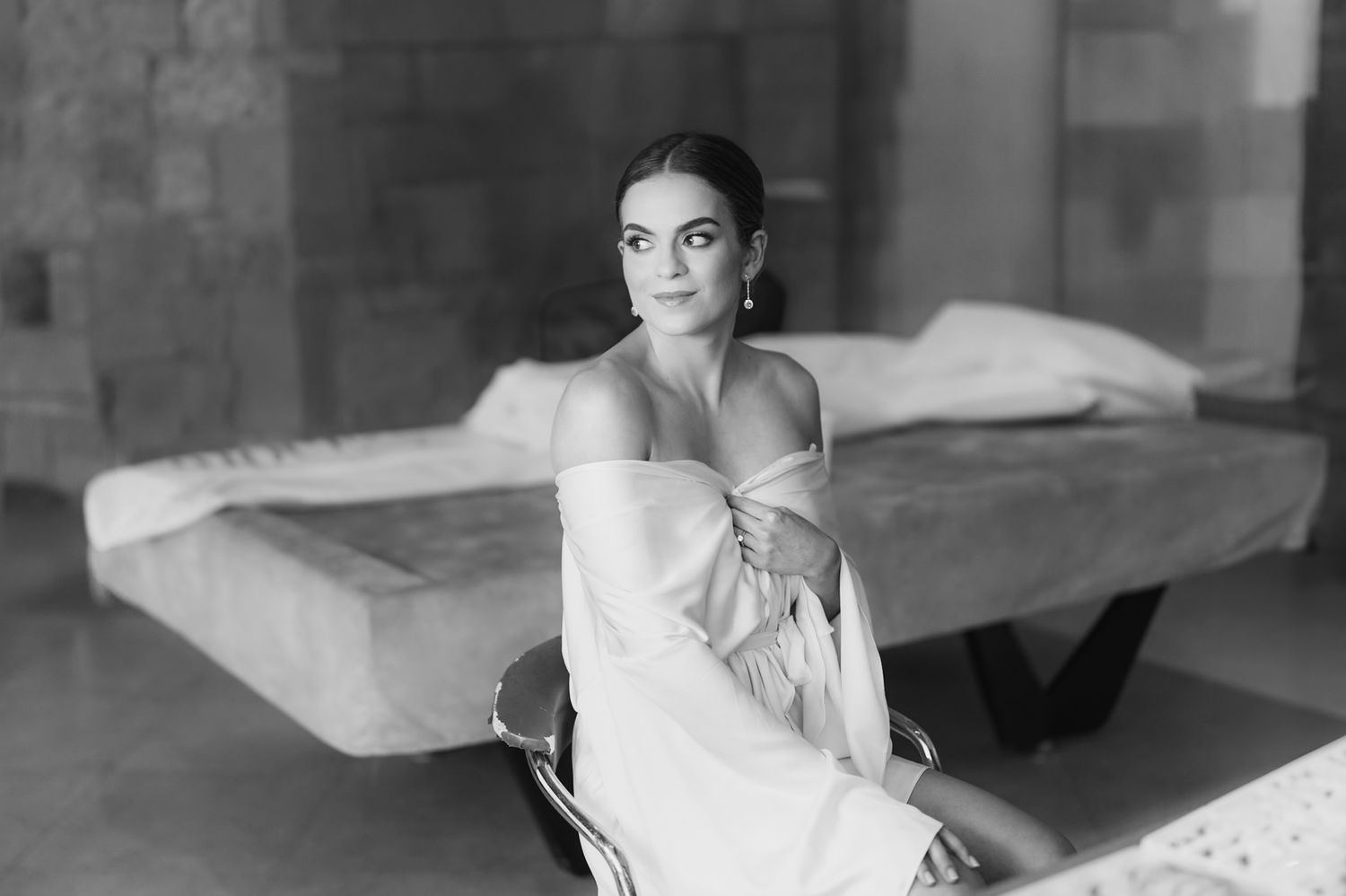Elegant black and white portrait of a bride in an off-shoulder robe seated in a sunlit stone room.