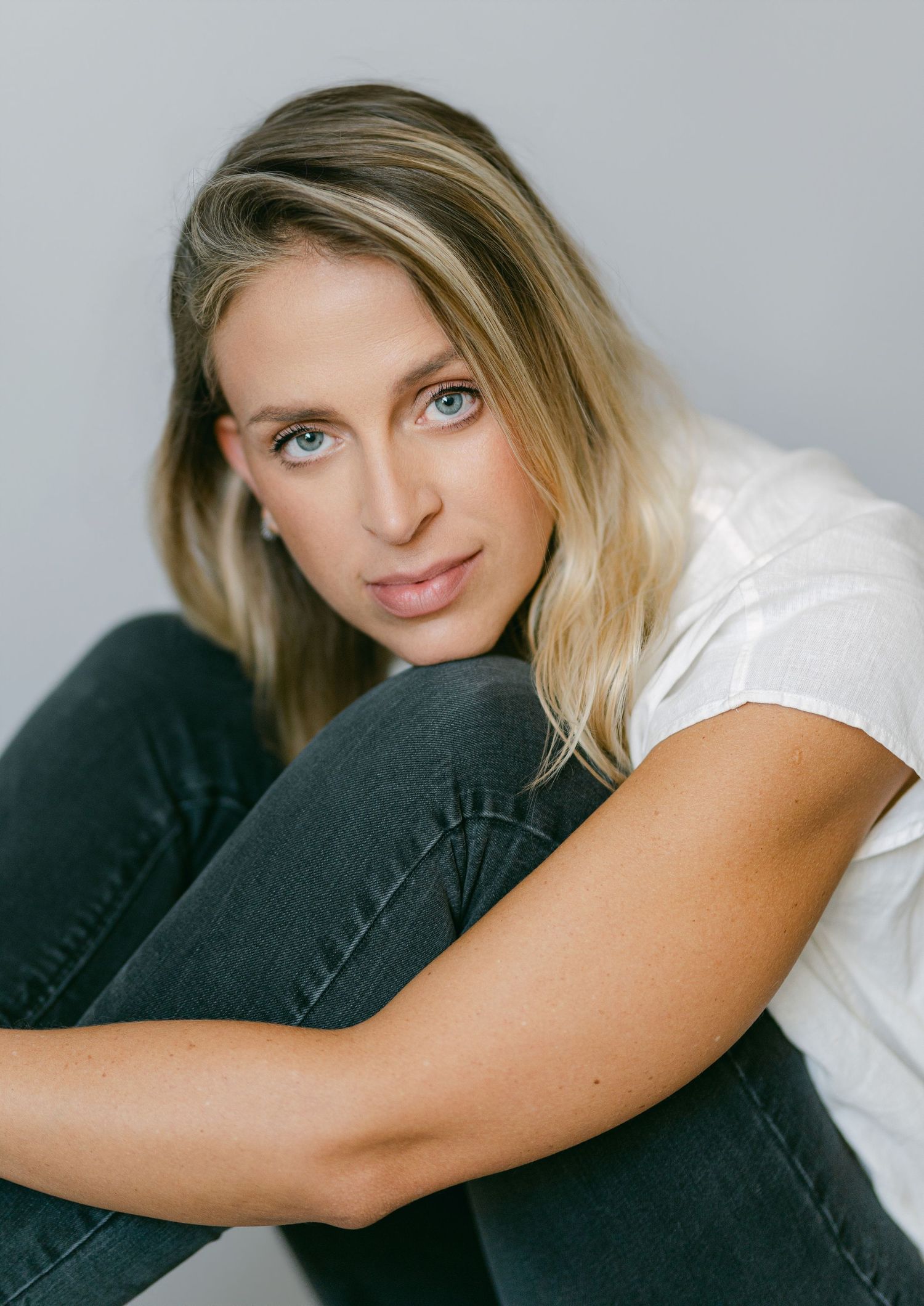 Blonde woman with blue eyes in white t-shirt and jeans sitting against gray wall, studio portrait.