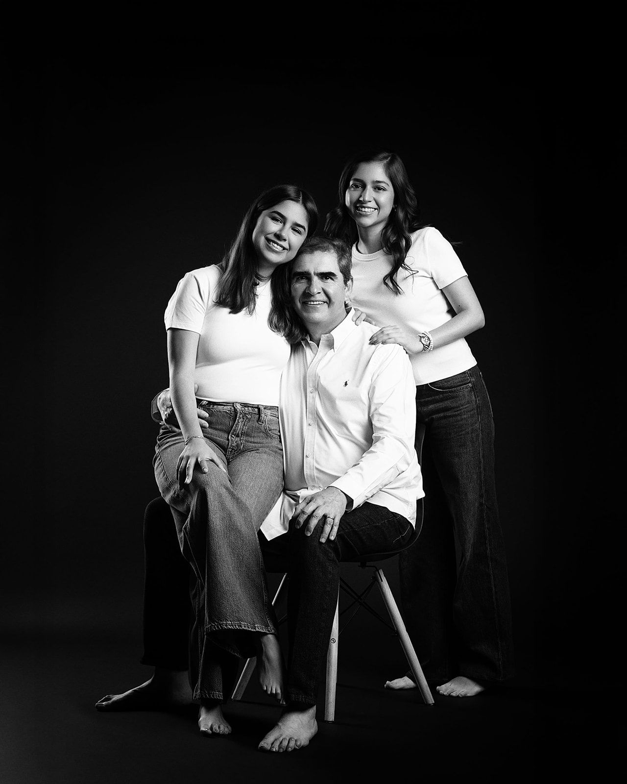 Black and white studio portrait of a father seated on a stool flanked by his two smiling daughters.