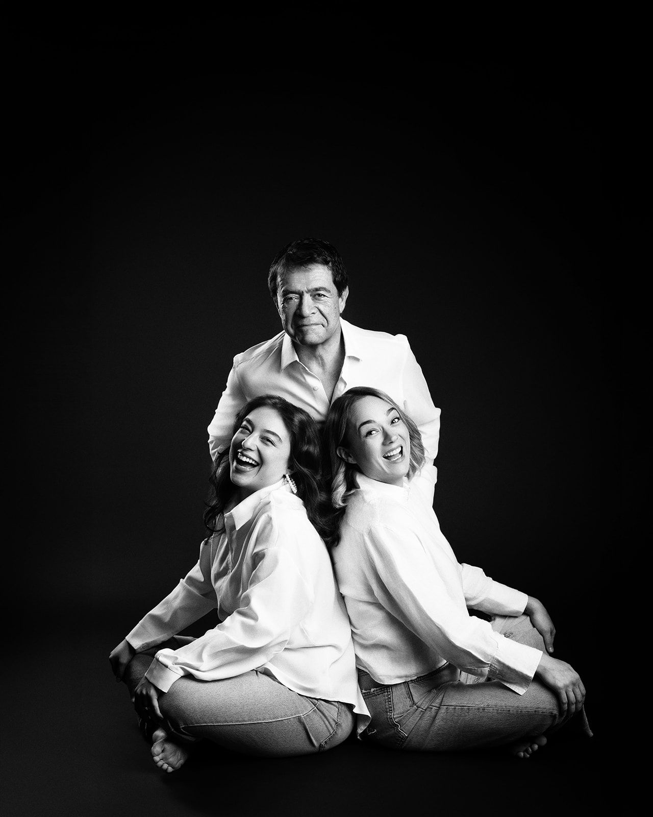 Black and white portrait of father and two laughing daughters posing on dark studio background.