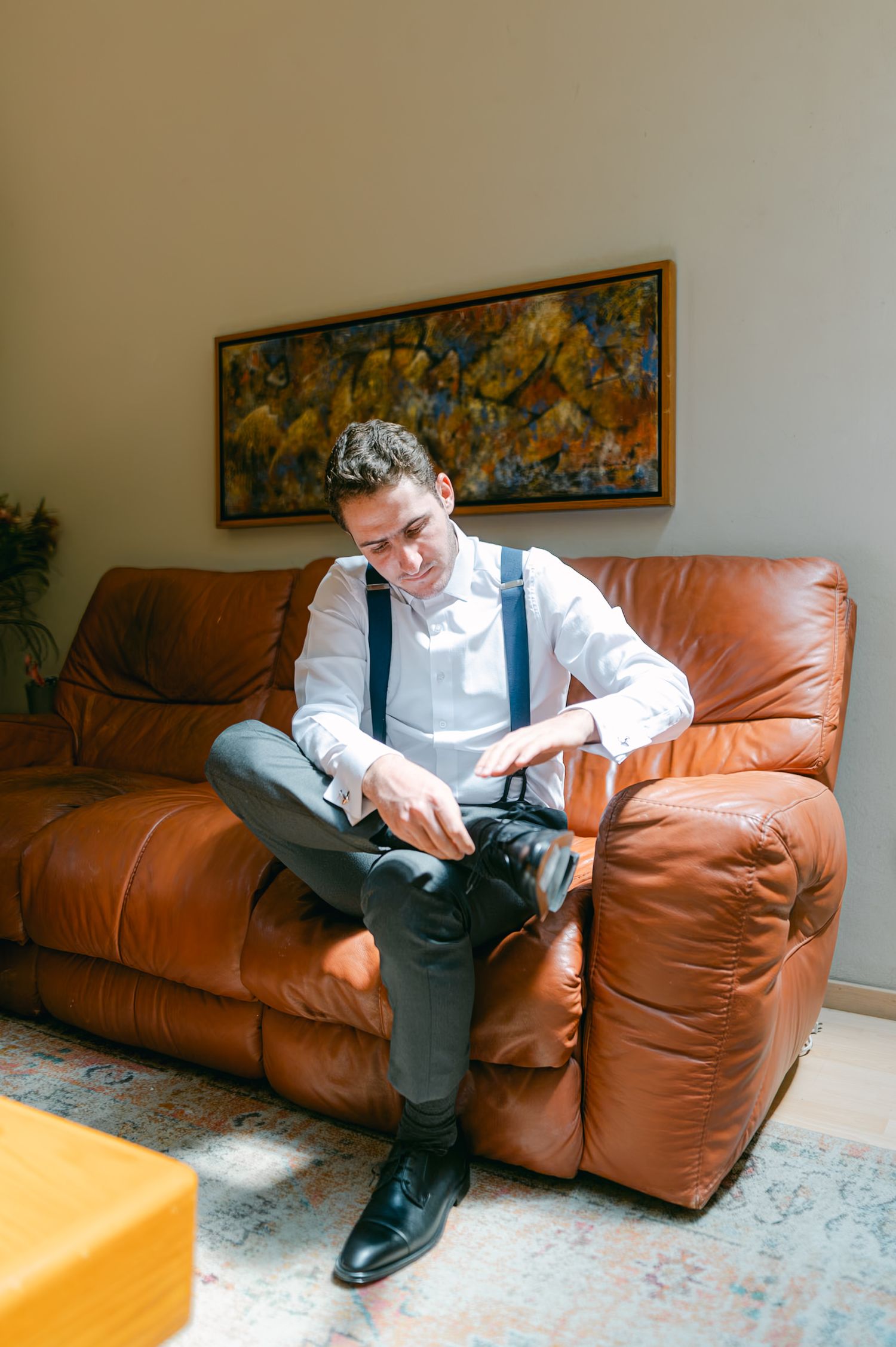 Groom sitting on brown leather sofa adjusting suspenders while getting ready for wedding day.