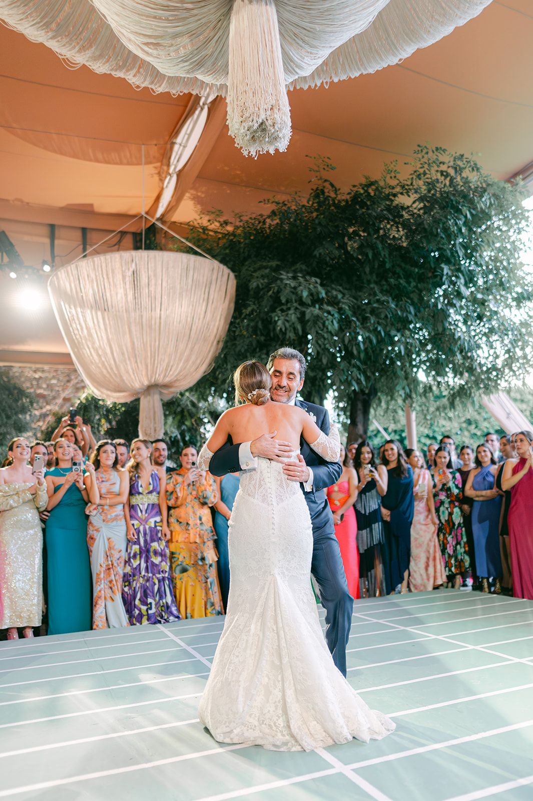 Newlyweds embrace during their first dance under a stunning chandelier with guests watching at a garden wedding.