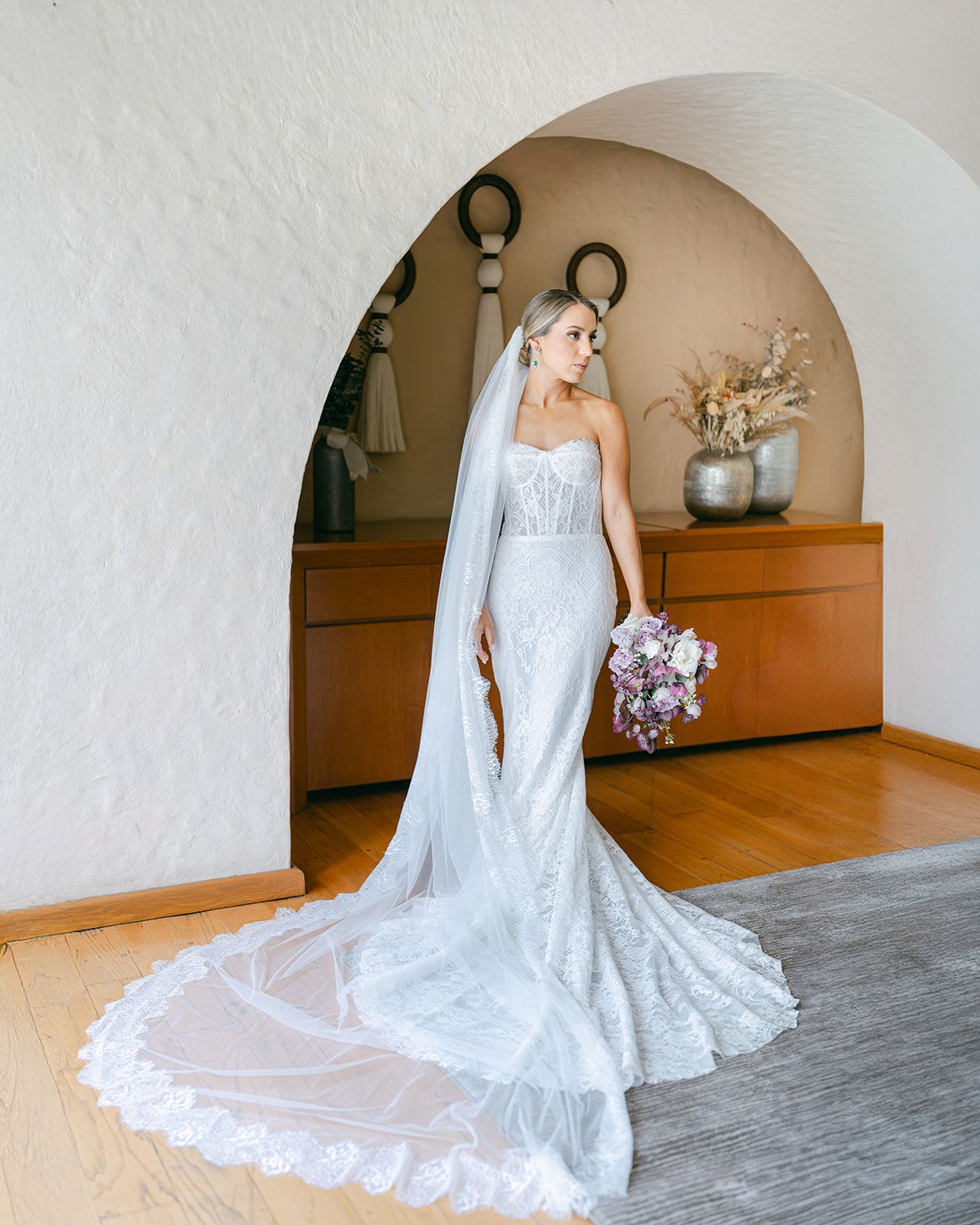 Bride in lace mermaid gown with cathedral veil holds purple bouquet under arch.