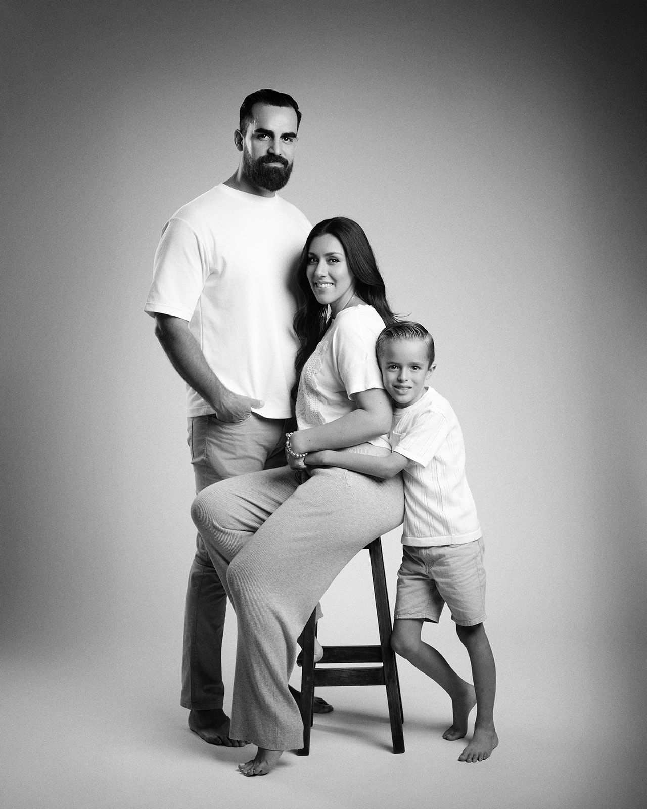 Black and white studio portrait of a bearded father, mother seated on a stool, and young son hugging her.