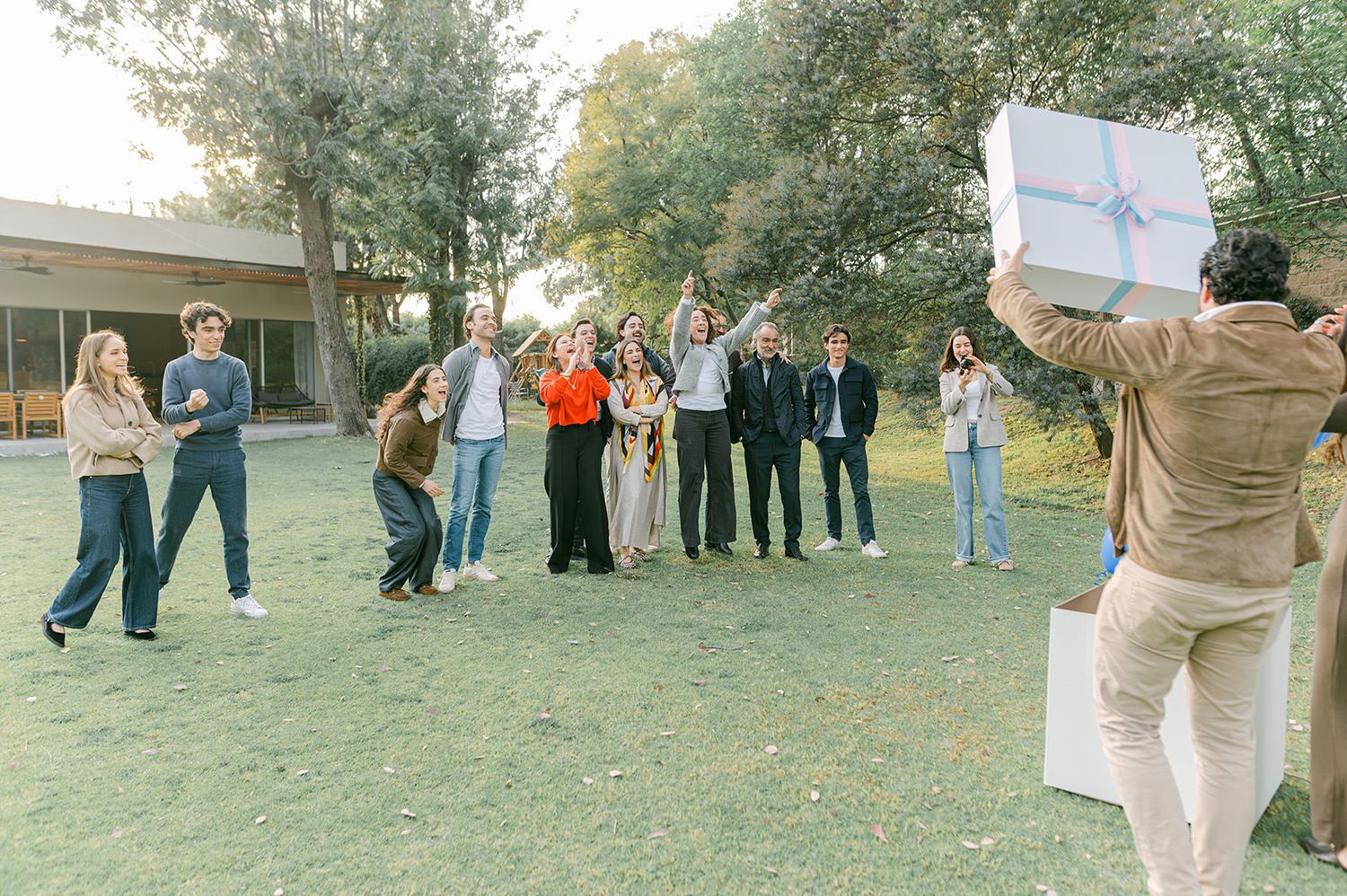 Group of friends celebrating gender reveal party on lawn with large gift box reveal.