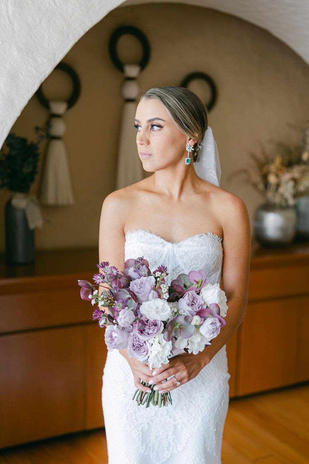 Bride in strapless lace gown holds purple and white floral bouquet near archway.