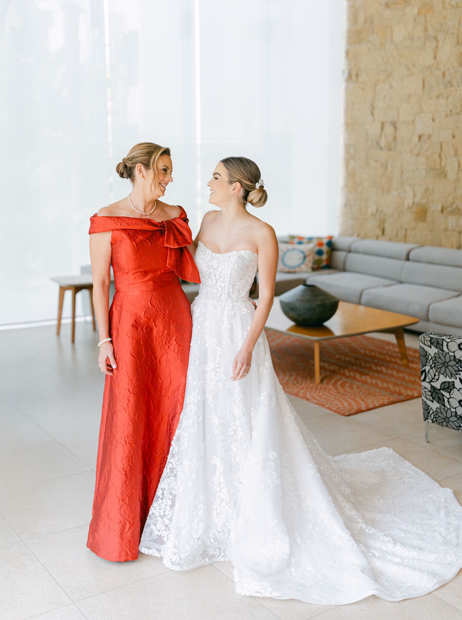 Bride in strapless lace gown sharing a tender moment with mother in red off-shoulder dress.