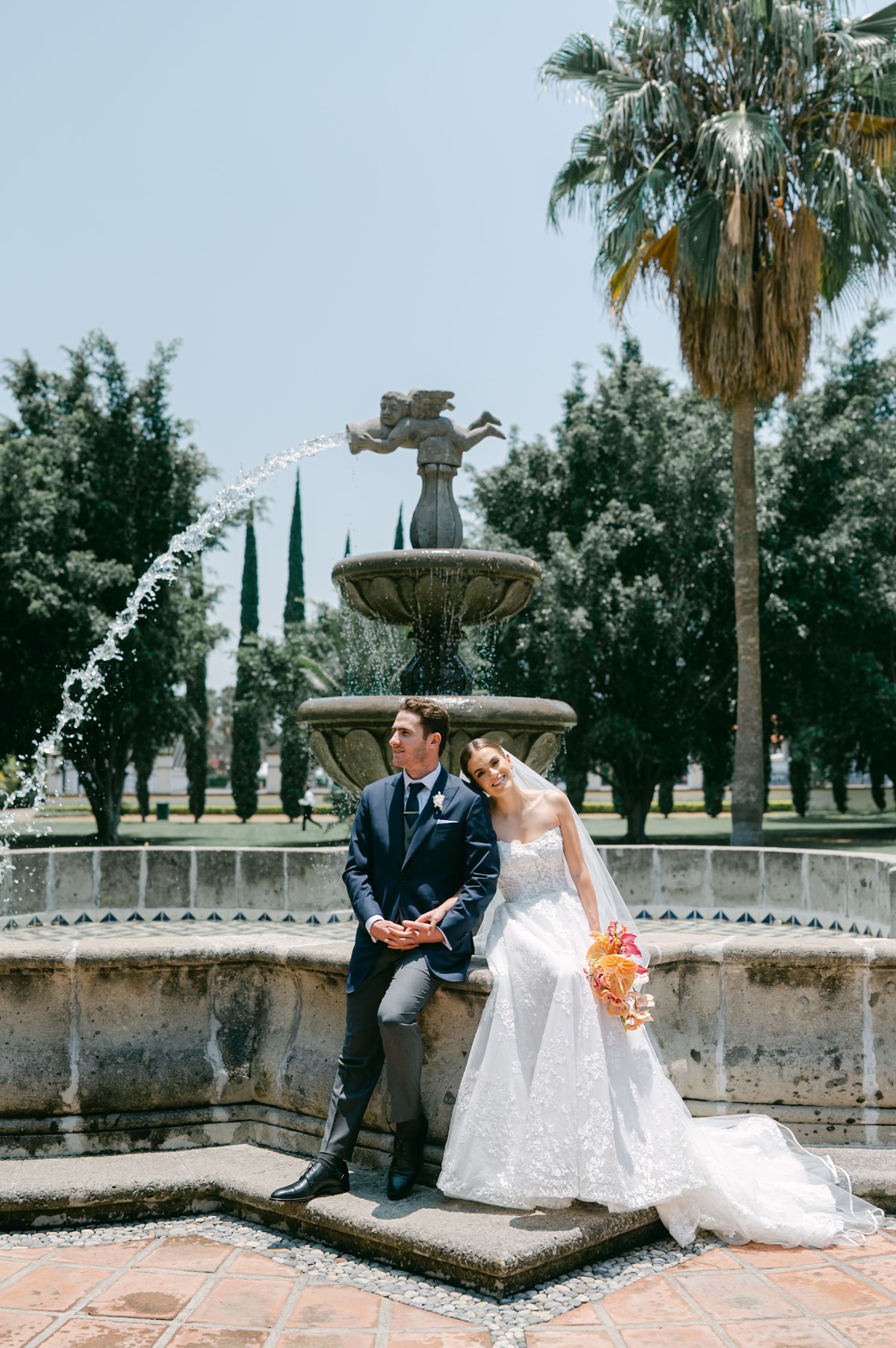 Bride and groom pose beside an ornate stone fountain with palm trees at outdoor wedding venue.