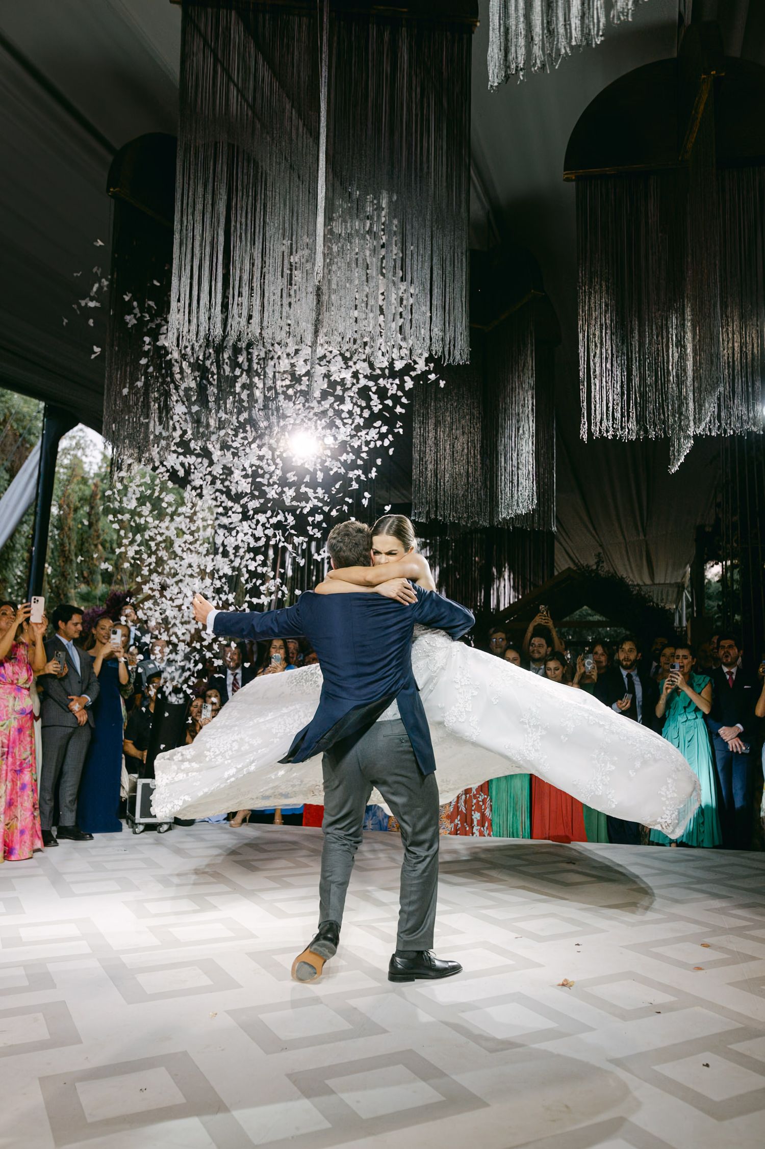 Groom lifts bride during dance as confetti explodes around them under hanging decor.