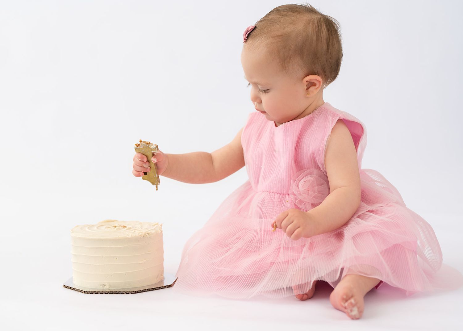 Baby girl in pink tutu dress reaches for gold candle on white birthday smash cake.