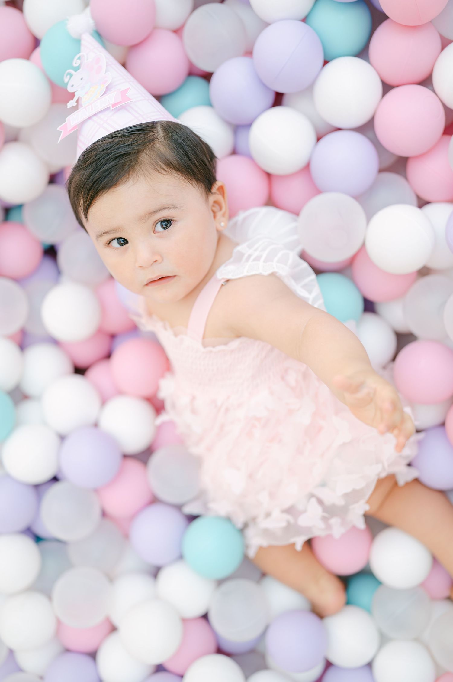 Adorable baby girl in a pink dress and party hat sitting in a colorful pastel ball pit.