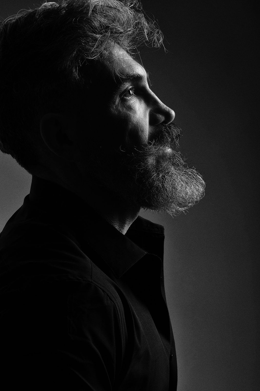 Black and white profile portrait of a bearded man with tousled hair, dramatic side lighting on dark background.
